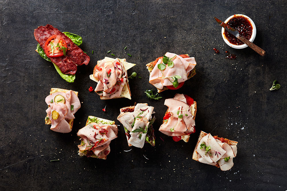 An assortment of open-faced sandwiches with sliced deli meat, greens, and vegetables on bread, arranged on a dark surface next to a strip of bacon and a small bowl of sauce.