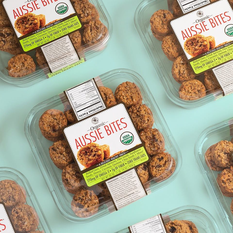 Plastic containers of Organic Aussie Bites are arranged on a light blue surface. The packaging displays product information and a USDA Organic label.