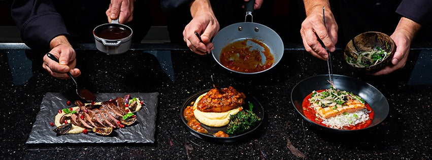 Three plates of gourmet dishes being garnished by chefs on a dark countertop: a beef dish, a chicken dish, and a seafood dish, each with various sauces and garnishes.