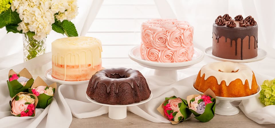 Five decorated cakes, including bundt and layered styles in chocolate, vanilla, and pink, displayed on stands with flower arrangements and cupcake bouquets on a white table.