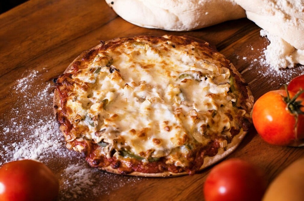 Bartolini's Sausage+Pepperoni+, Mushroom Thin Crust Pizza features melted cheese, savory sausage, and pepperoni on a crispy crust, shown atop a wooden surface with flour, tomatoes, and dough in the background.