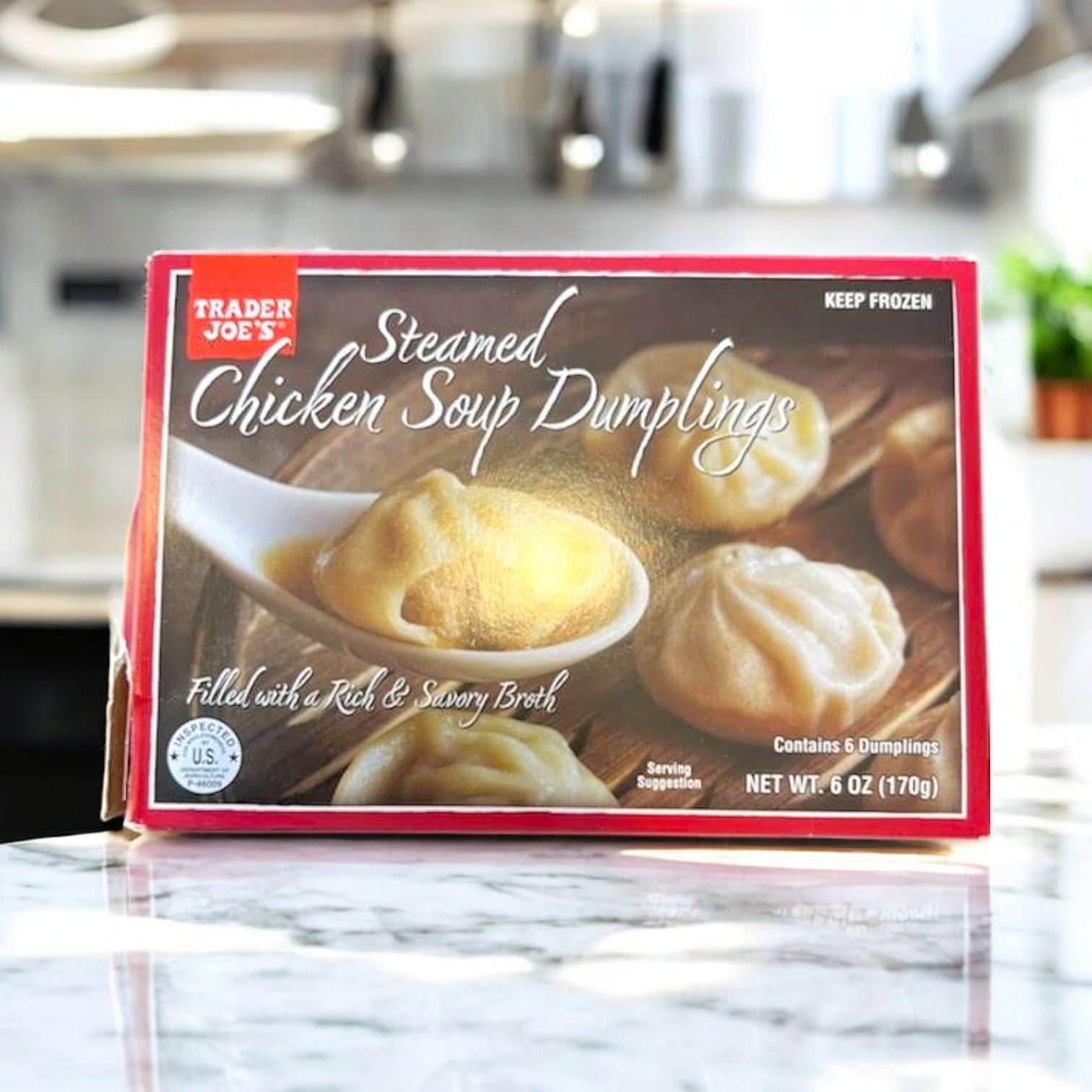 A Easy Lunches Steamed Chicken Soup Dumplings box, 6 oz, lies on a marble counter, displaying dumplings in a spoon. The packaging emphasizes weight and serving details for freshness. Enhance this versatile dish with julienned green pepper for added flavor.