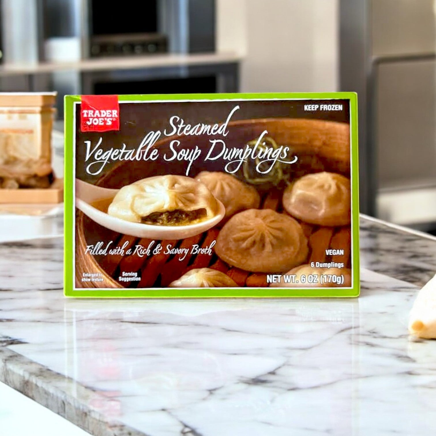 A box of Easy Lunches Steamed Vegetable Soup Dumplings – 6 oz. rests on a kitchen counter, highlighting their vegan and plant-based appeal, with packaging displaying these tempting dumplings filled with a savory broth inspired by Asian cuisine.