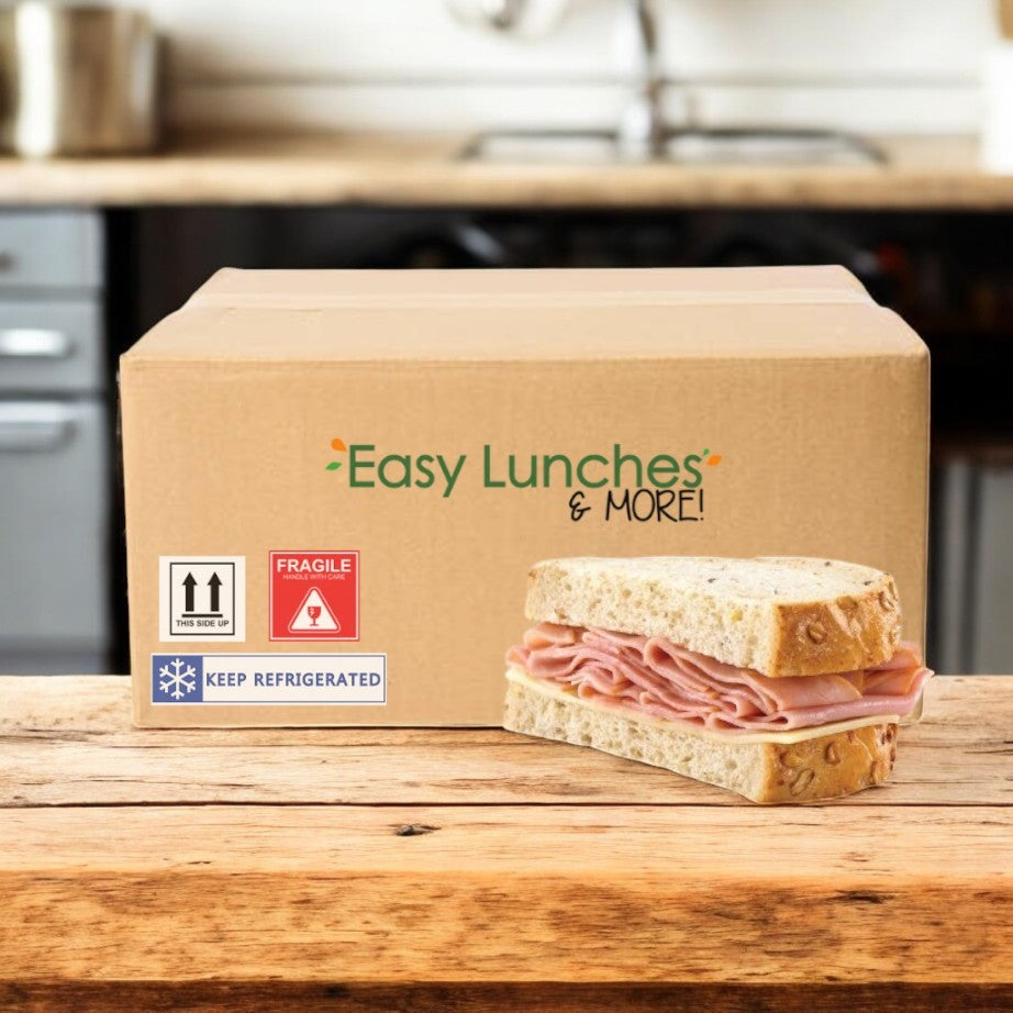 A cardboard box labeled "Easy Lunches & More!" from E.A. Sween sits on a counter with a Ham & Swiss sandwich on oat-topped wheat bread in front. Stickers show it's fragile and must be refrigerated; ships frozen, lasts 30 days in cooler.