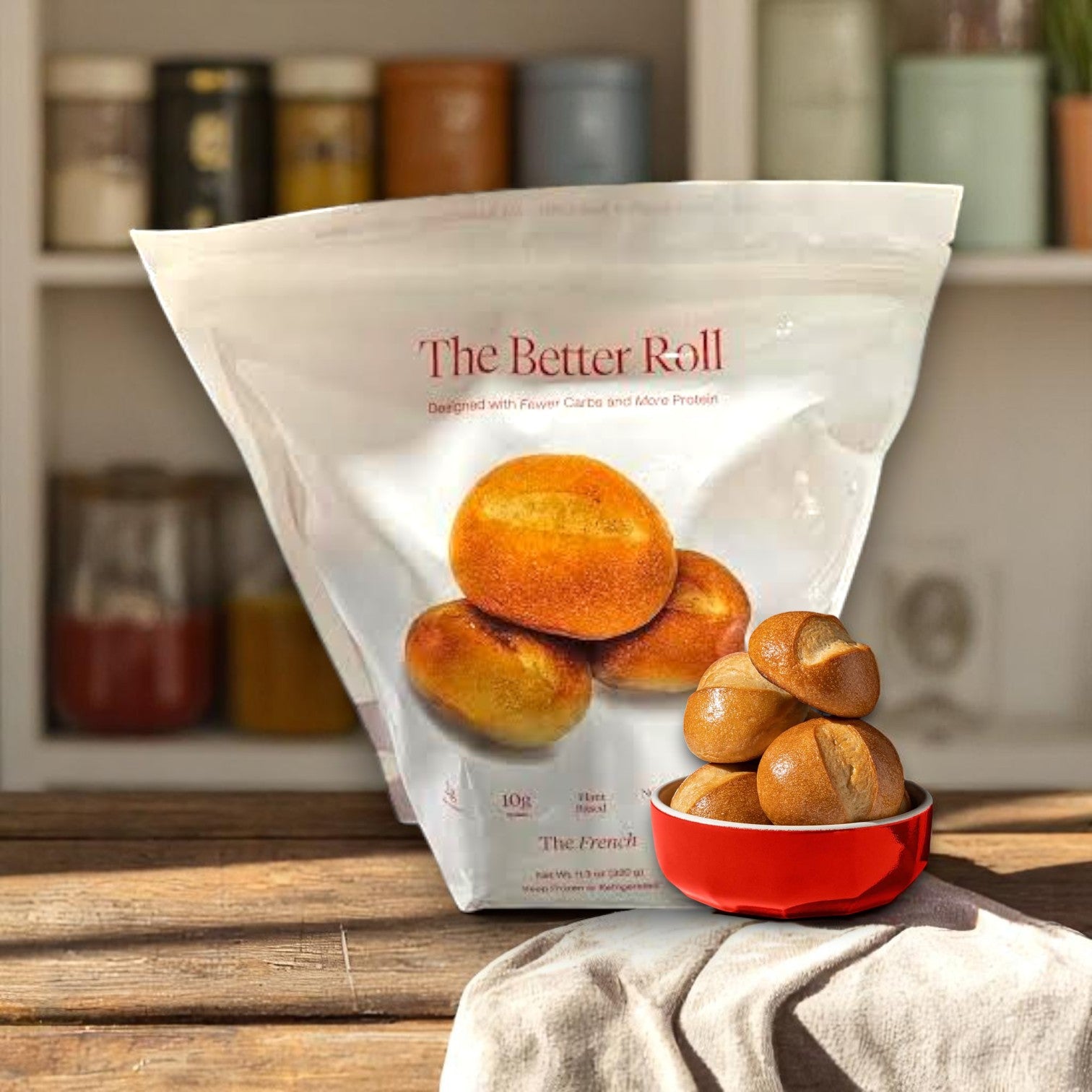 A bag of BetterBrand's The Better Roll – French Style (8 rolls) is behind a red bowl filled with artisan-style rolls, set on a kitchen table. The product is in stock and ready to ship!.