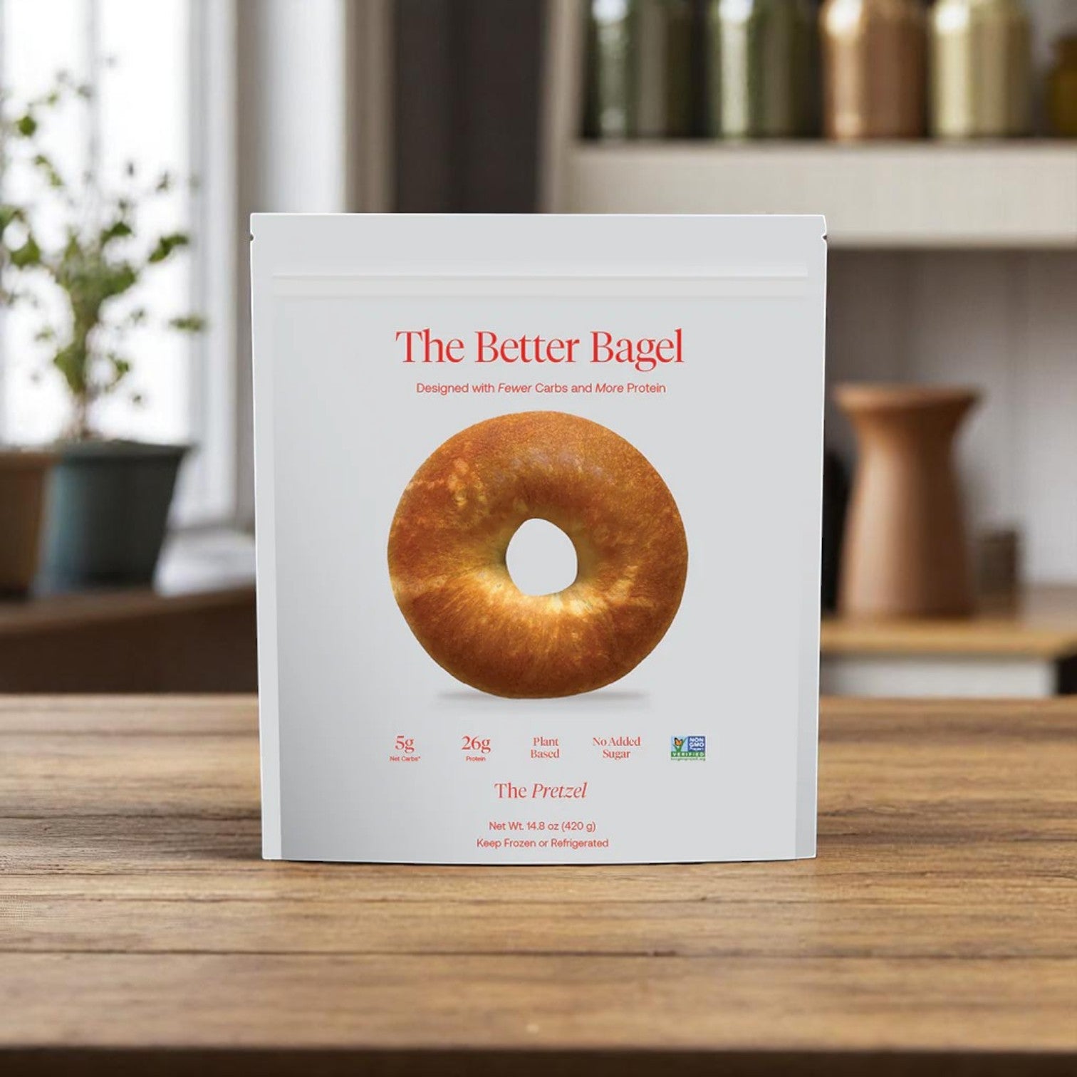 A white BetterBrand bag labeled "The Better Bagel – Pretzel Style" (1 bag, 4 bagels) sits on a wooden kitchen counter, with blurred kitchen items in the background. In stock and ready to ship!.