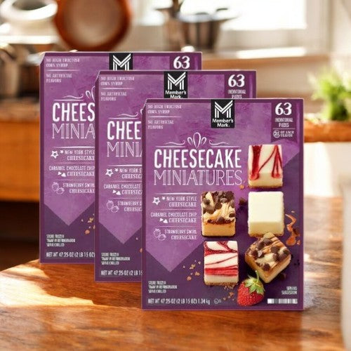 Three boxes of Member's Mark Cheesecake Mini's Variety Pack are displayed on a wooden surface, offering indulgent bite-sized cheesecake flavors. Perfect for any sweet tooth!.
