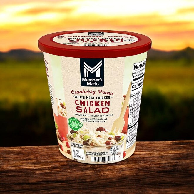A 2 lb container of Member's Mark Cranberry Pecan Chicken Salad sits on a wooden surface, with an outdoor landscape in the background.