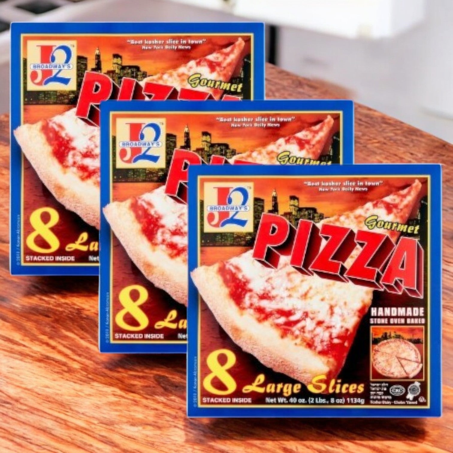 Three boxes of J2L Broadway's Gourmet Pizza, branded as "J2 Broadway's Large Slices Gourmet Pizza-3 Count," are stacked on a wooden surface. Each box holds eight large slices known for bold flavors.
