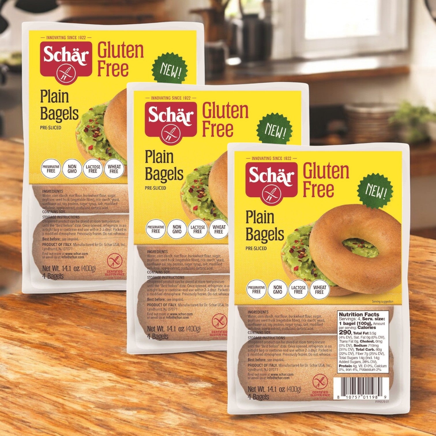 Three packages of Schar Gluten Free Plain Bagel, 14.1 oz. - 3 Count are on a wooden surface with a kitchen background. Each package highlights the classic taste and shows an image of a bagel topped with avocado spread.