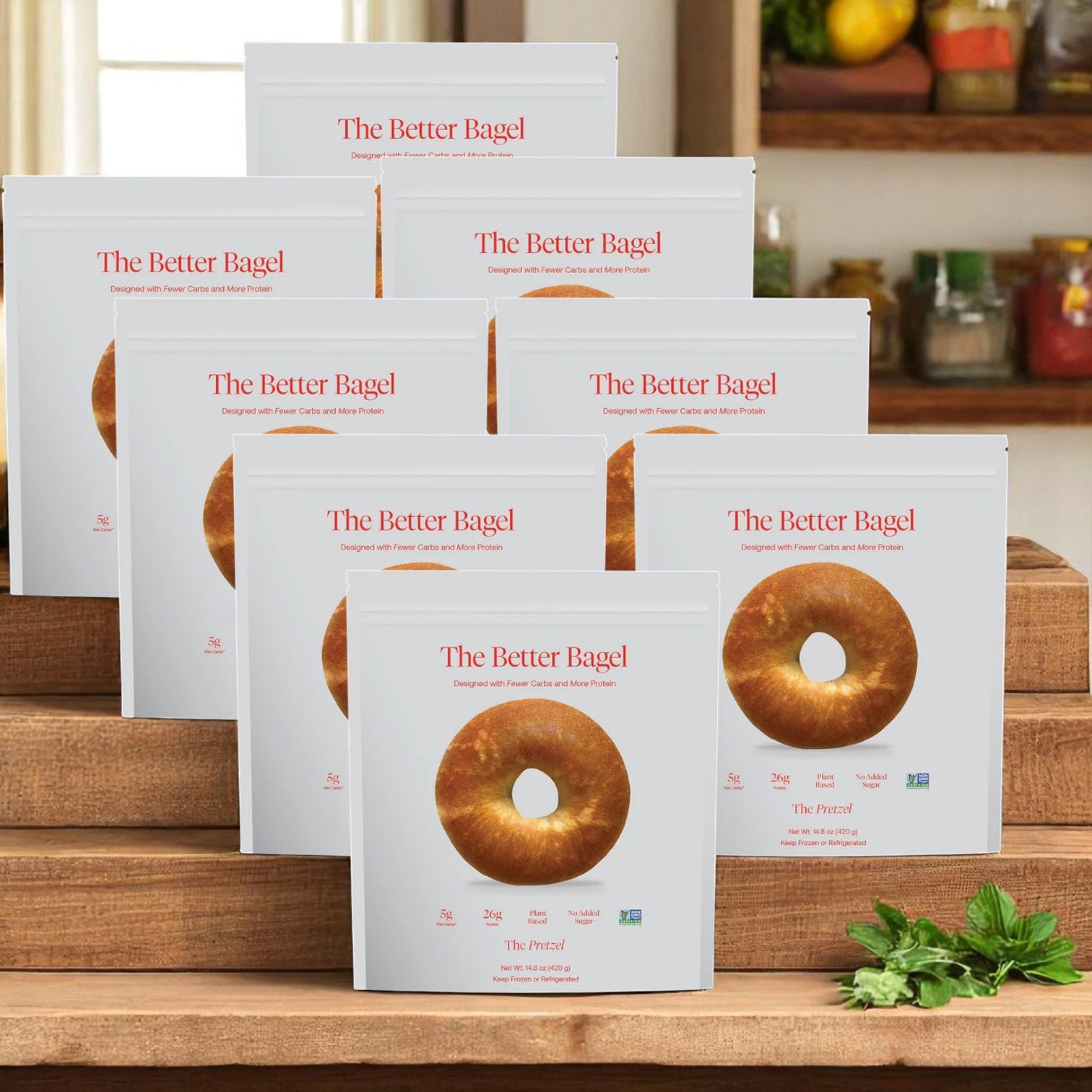 Boxes of BetterBrand’s The Better Bagel – Pretzel Style (8 bags, 32 bagels total) are displayed on wooden steps in a kitchen, with herbs and jars in the background. In stock and ready to ship!.