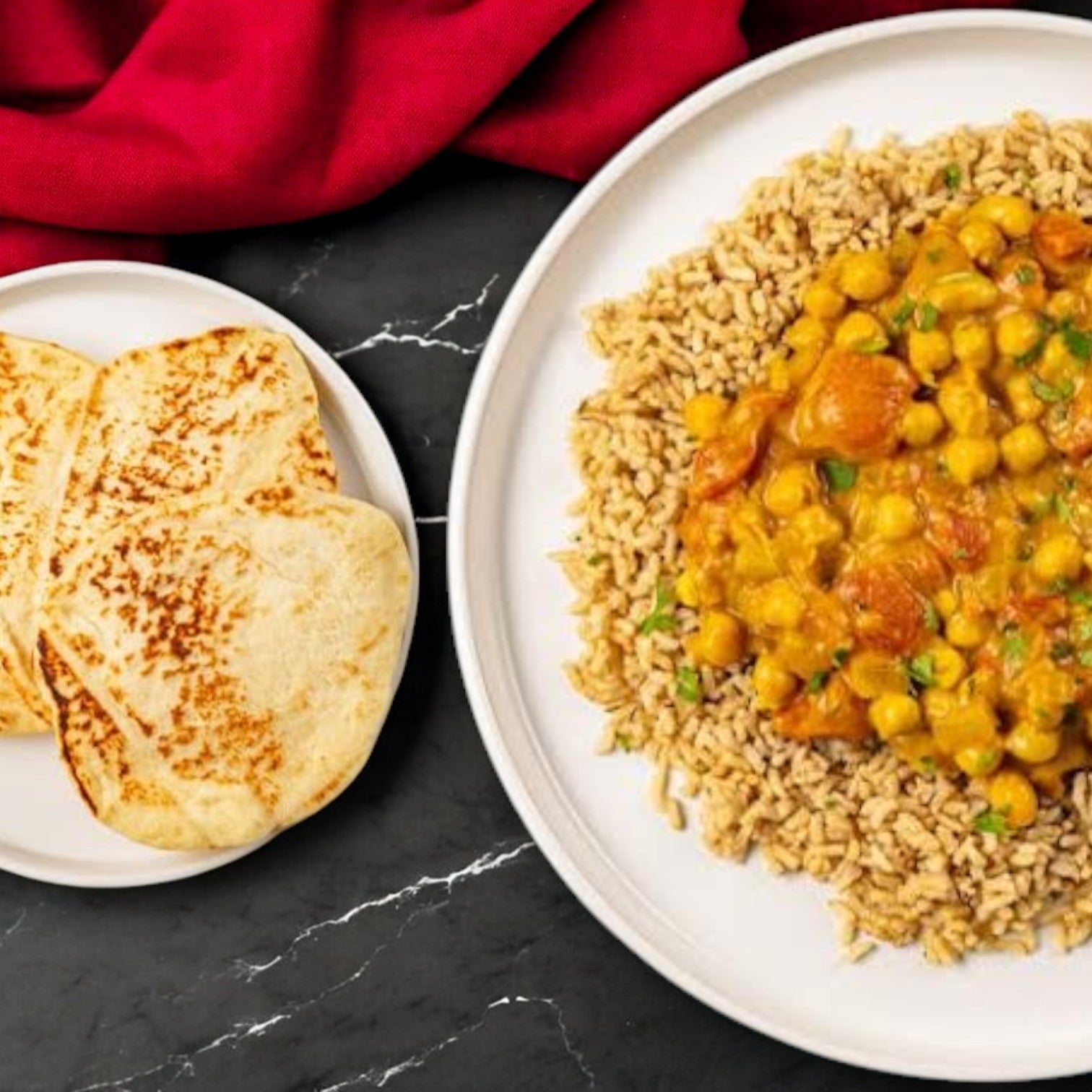 A plate of rice topped with FoodHaul's Chana Masala by Jay Farley, a staple in Indian cuisine, and a side of naan bread rests on a dark marble surface with a red cloth nearby.