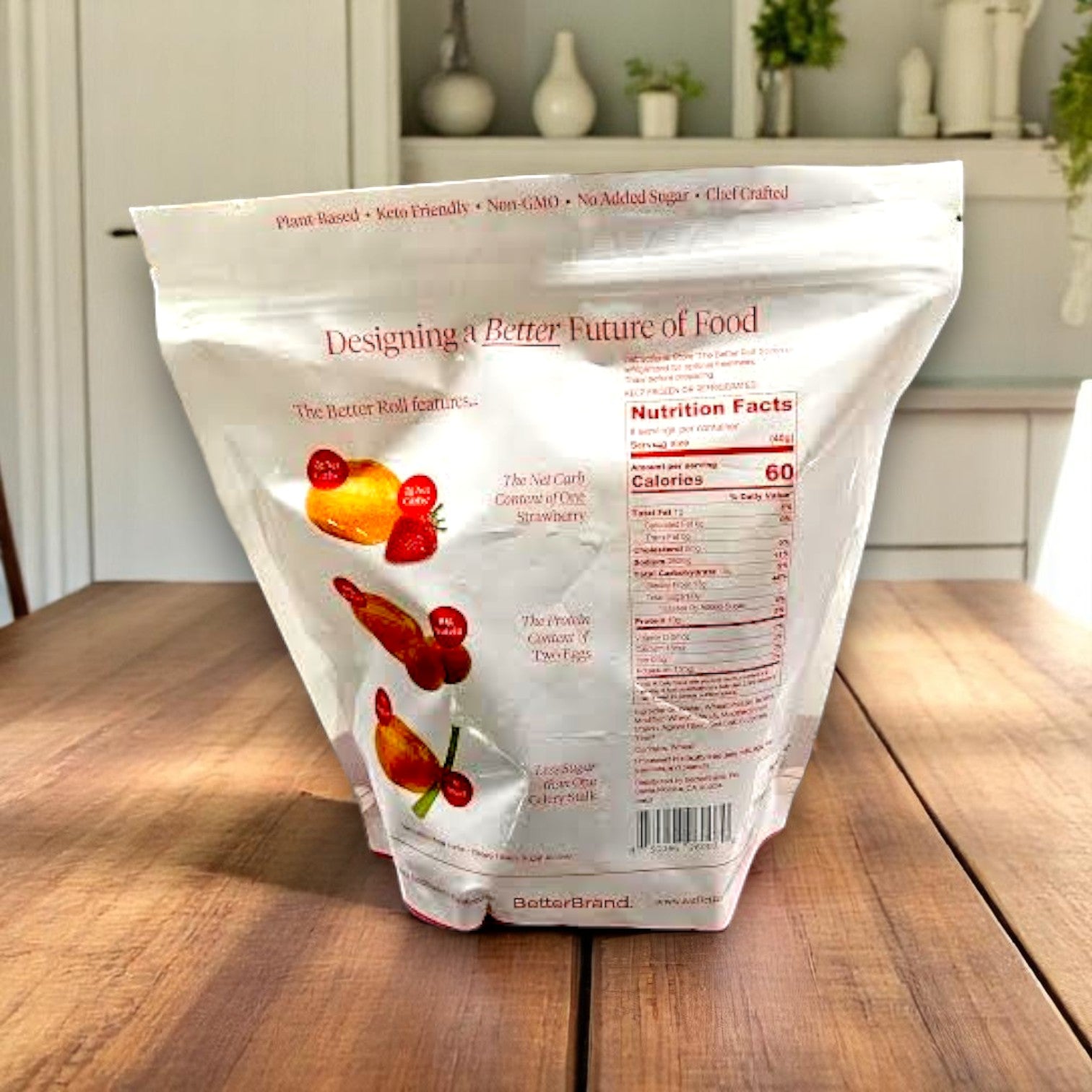 A large white resealable bag labeled "BetterBrand" sits on a wooden table, featuring The Better Roll – French Style (8 Rolls per Bag). Kitchen shelves and plants create a cozy backdrop, highlighting its artisan-style appeal. In stock and ready to ship!.