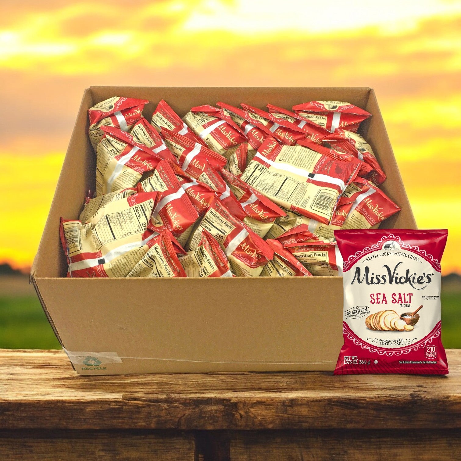A box of Miss Vickie’s Chip Potato Regular Sea Salt 64/1.375 oz. bags sits on a wooden surface with a sunset in the background.