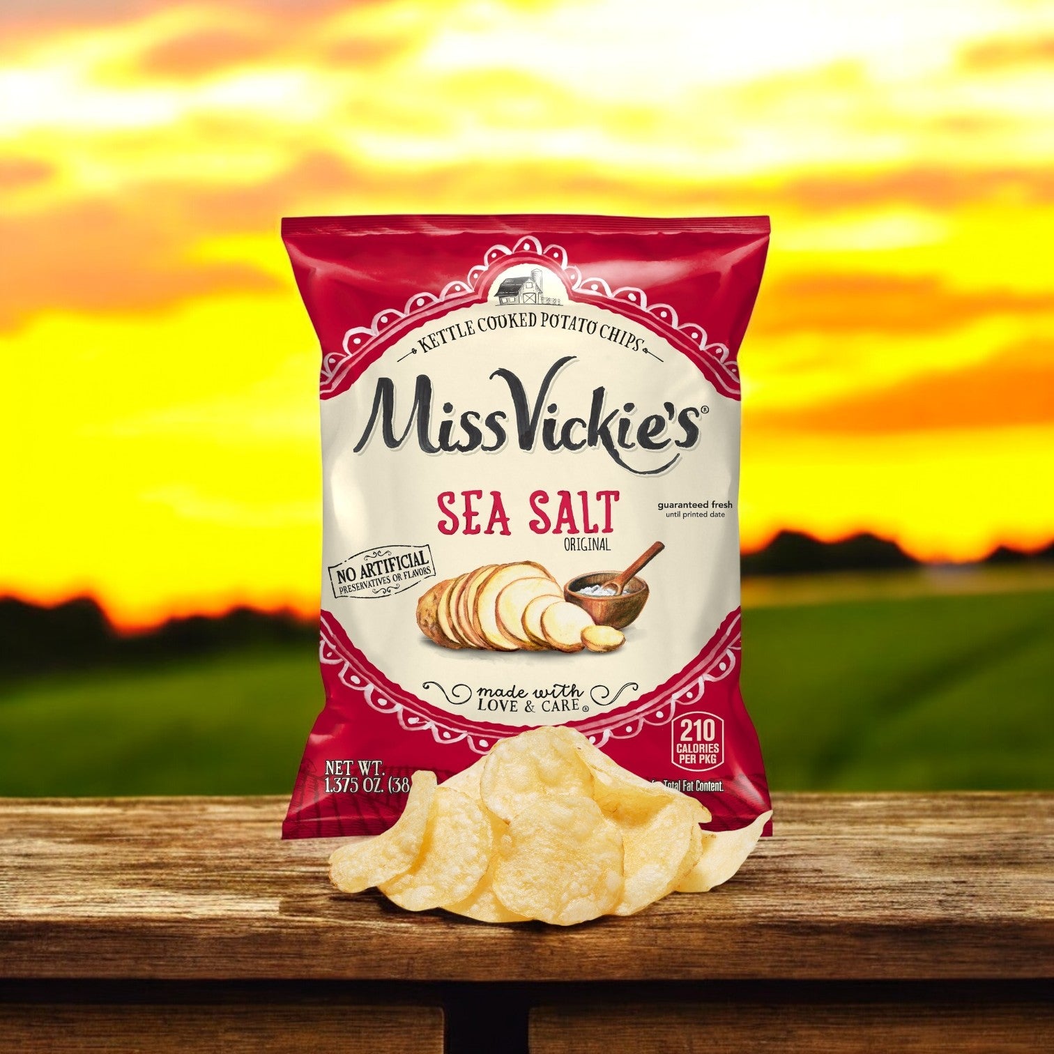 A bag of Miss Vickie's Chip Potato Regular Sea Salt 64/1.375 oz. sits on a wooden surface outdoors, golden chips in front and a sunset field in the background.