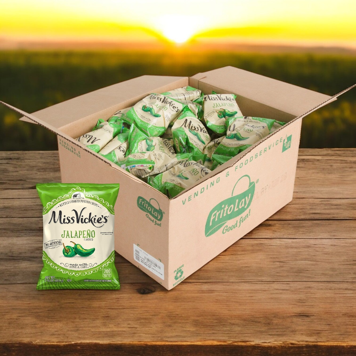 A box of Miss Vickie’s Chip Potato Jalapeño Kettle 64/1.375 oz. sits on a wooden table outdoors at sunset, with one spicy Miss Vickie's snack bag displayed in front.