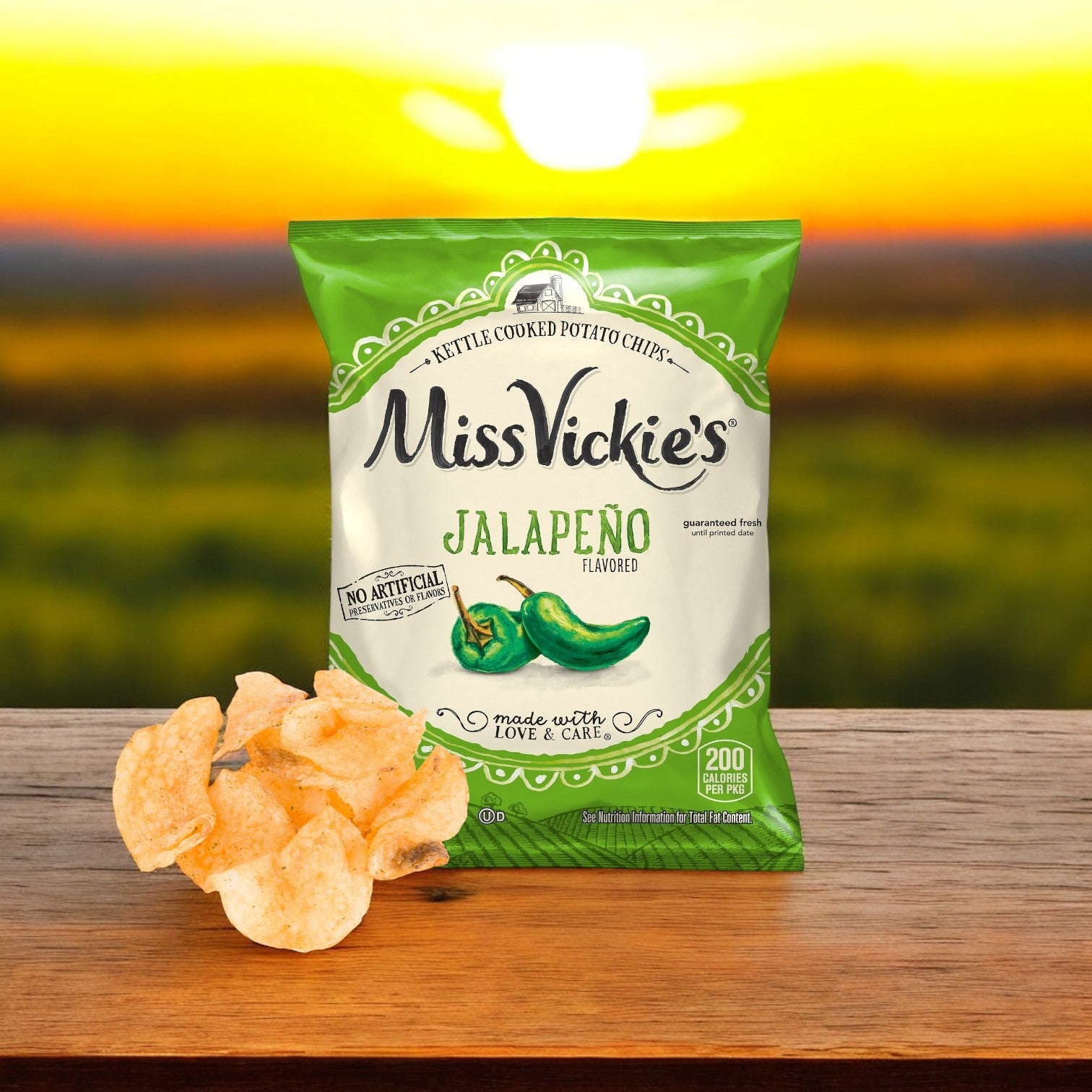 A bag of Miss Vickie’s Chip Potato Jalapeño Kettle (64/1.375 oz.) sits on a wooden surface with crunchy kettle chips beside it, set against a blurred outdoor sunset for the perfect spicy snack moment.