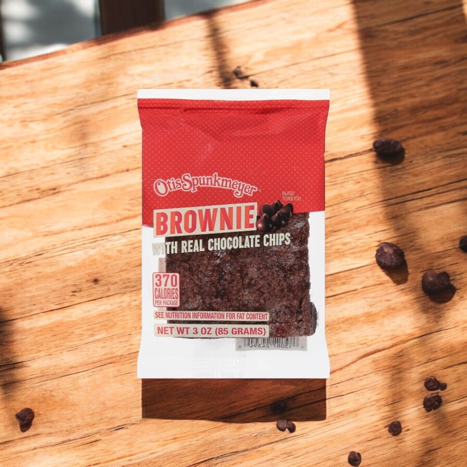 A 3-ounce Otis Spunkmeyer brownie, bursting with real chocolate chips, lies individually wrapped on a wooden surface as sunlight creates charming shadows.