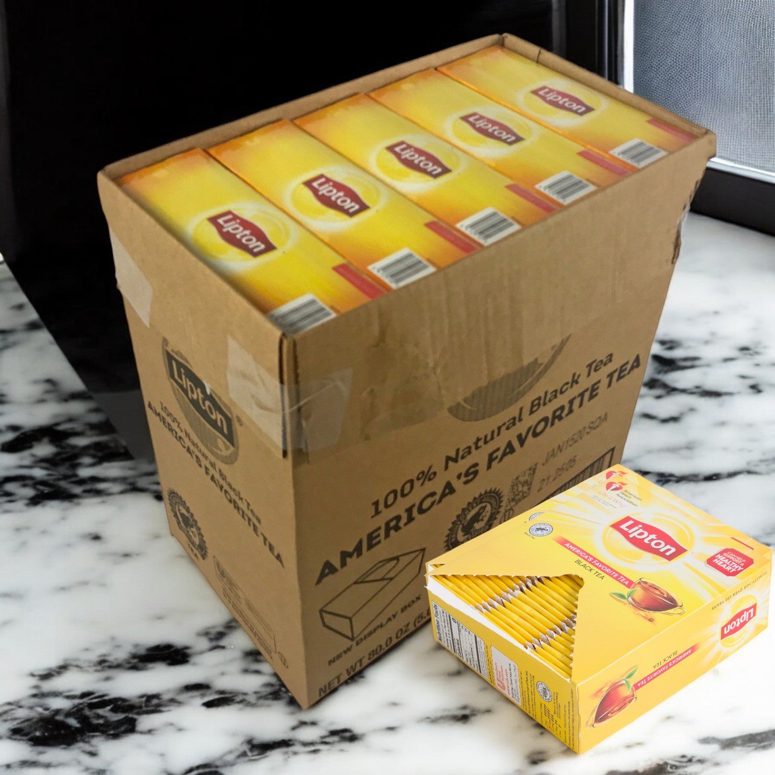 An open cardboard box of Lipton Tea Hot Bag Black Envelope 10/100 CT reveals several yellow boxes with individually wrapped tea bags inside; a separate Lipton black tea box is on the marble counter beside it.