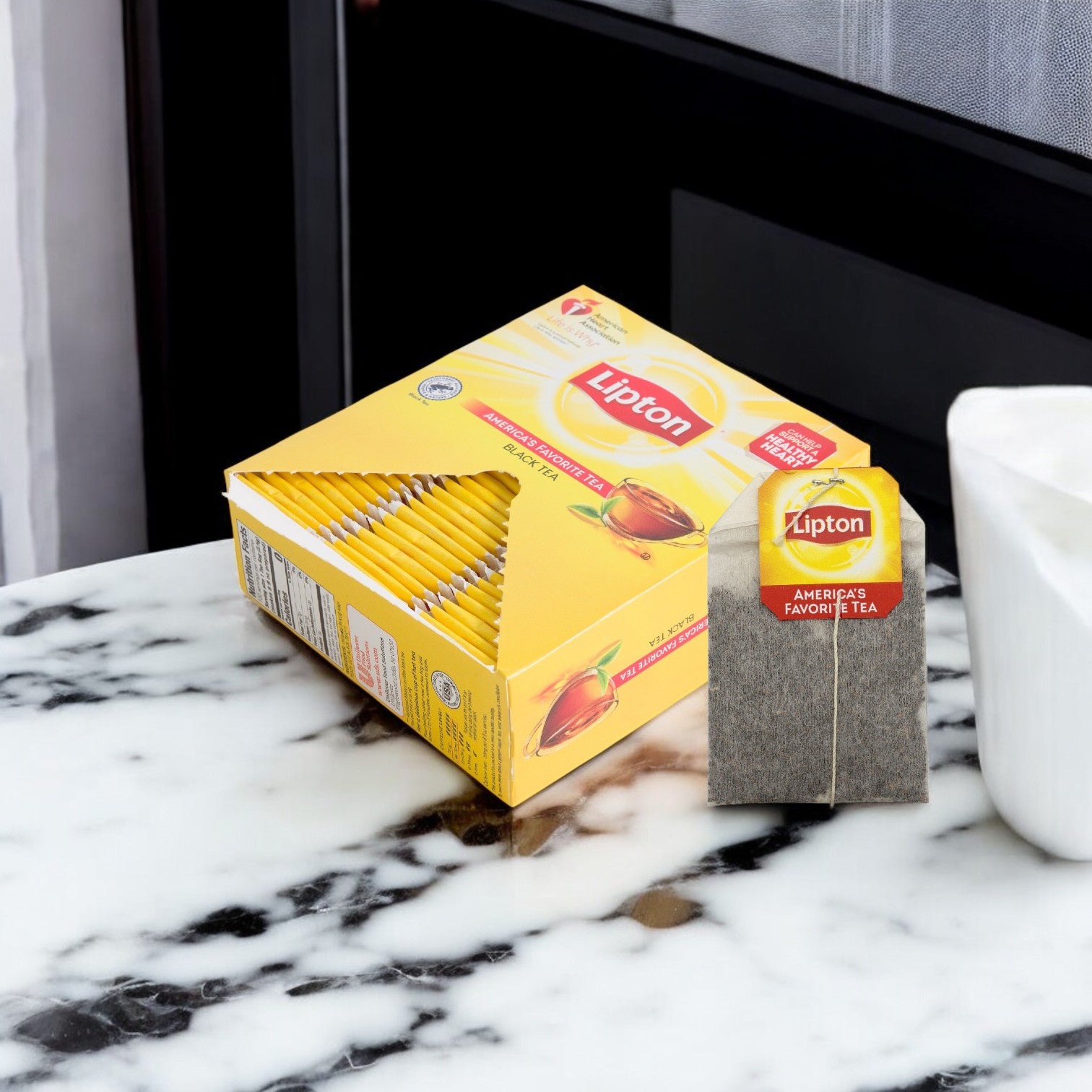 A box of Lipton Tea Hot Bag Black Envelope 10/100 CT sits on a marble countertop beside an individually wrapped Lipton tea bag and a white ceramic container.