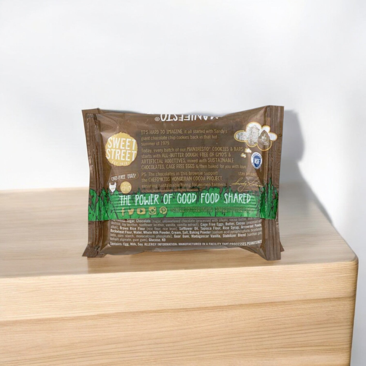 A Sweet Street Honduran Chocolate Manifesto Brownie, 3 oz., from a 48-count case, made with premium rich Honduran chocolate, sits on a wooden surface with its back showing the ingredients and product details.