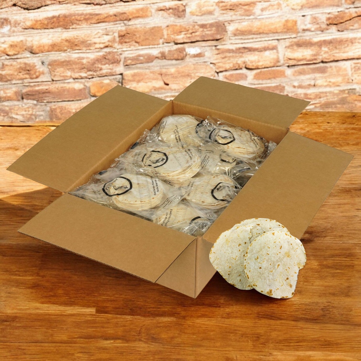 An open box on a wooden surface displays a bulk pack of El Milagro White Flour Tortillas, 5 inches each, individually wrapped for freshness. Three tortillas lie unwrapped next to the box against a backdrop of rustic brick.