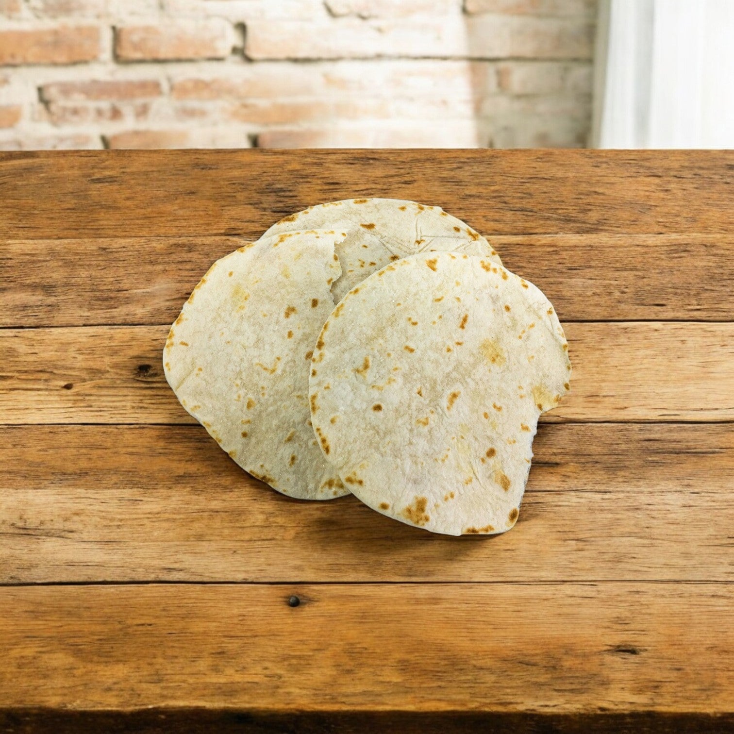 Two 5-inch corn tortillas on a wooden table with a brick wall behind evoke the quality of El Milagro's White Flour Tortillas (5-Inch - 42 Packs of 1 Dozen).