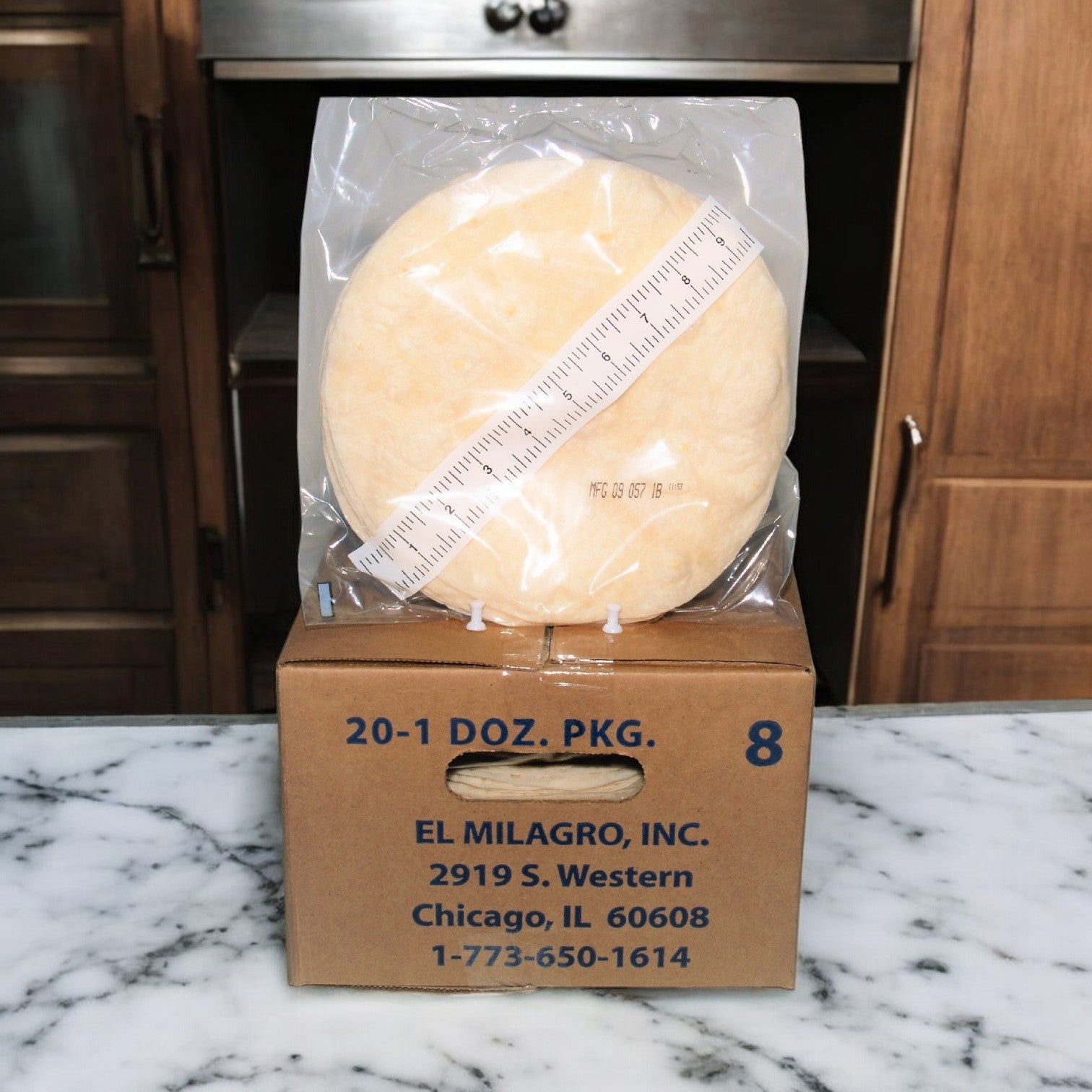 An El Milagro Flour Tortillas package—8 inches, 20 packs of a dozen—rests in a sealed plastic bag on a branded cardboard box on a marble counter with a ruler alongside.