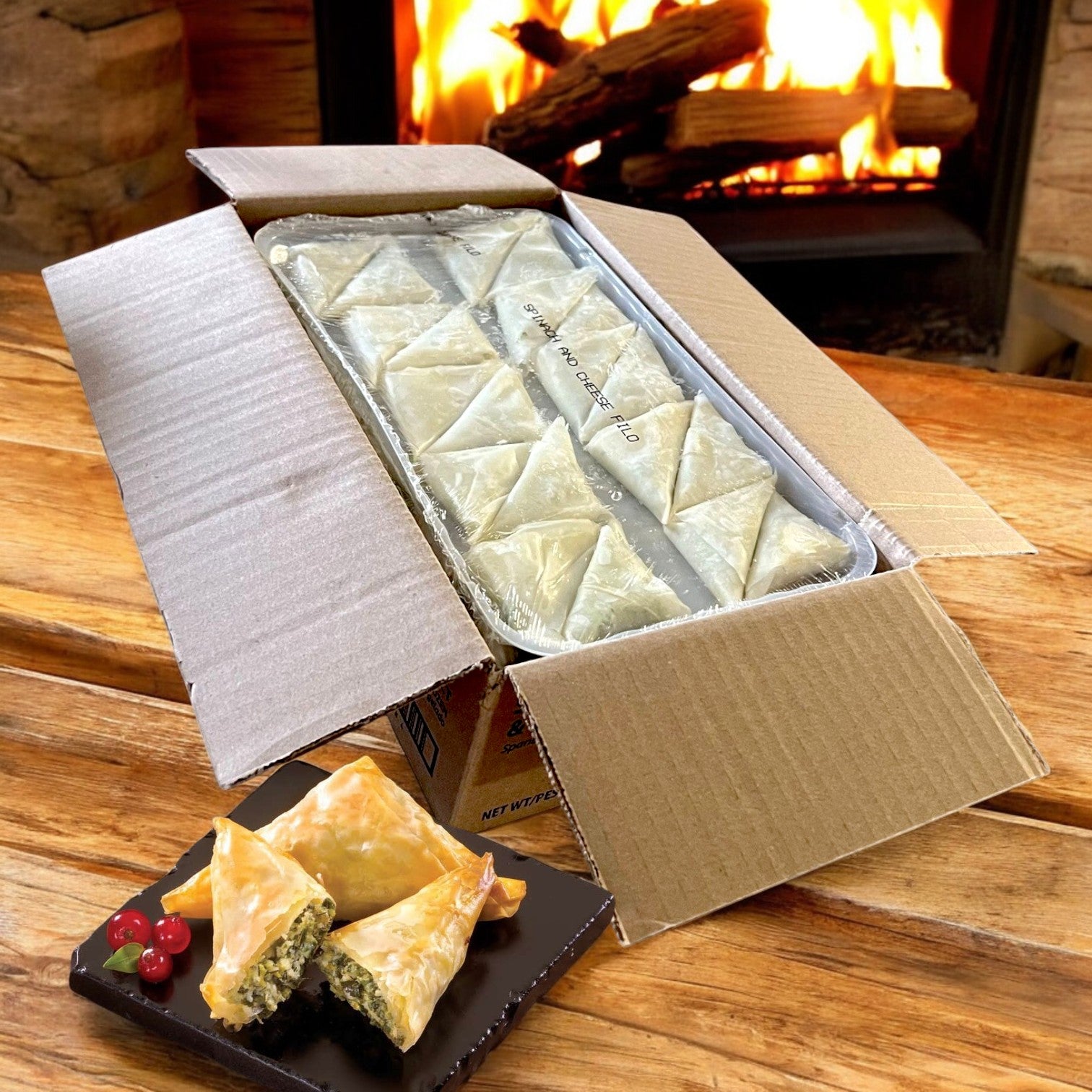 A box of Portico Classic Seafood Sysco Imperial Spanakopita Appetizer (5 trays of 20 count, frozen) sits on a wooden table beside a plate of flaky phyllo triangles with garnish and a fireplace in the background.