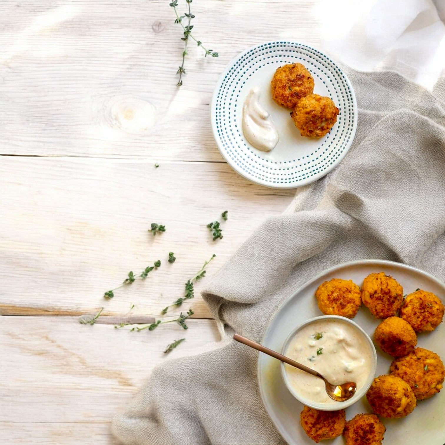 Two plates featuring Sysco No Fork Required Mini Crab Cakes – 100 Count, made with real crab meat, are served with creamy herbed dipping sauce on a light wooden surface and a beige cloth.