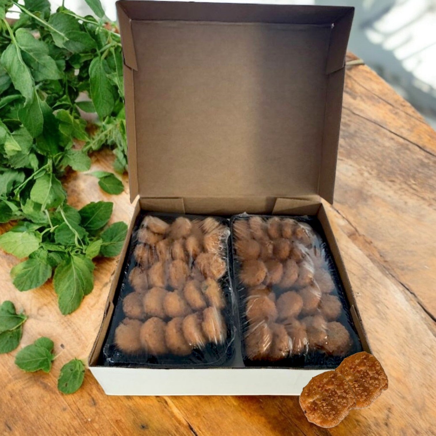 A Sysco No Fork Required Mini Crab Cakes – 100 Count box sits open on a wooden surface, revealing two plastic-wrapped trays of real crab meat mini cakes; mint leaves are next to the box.