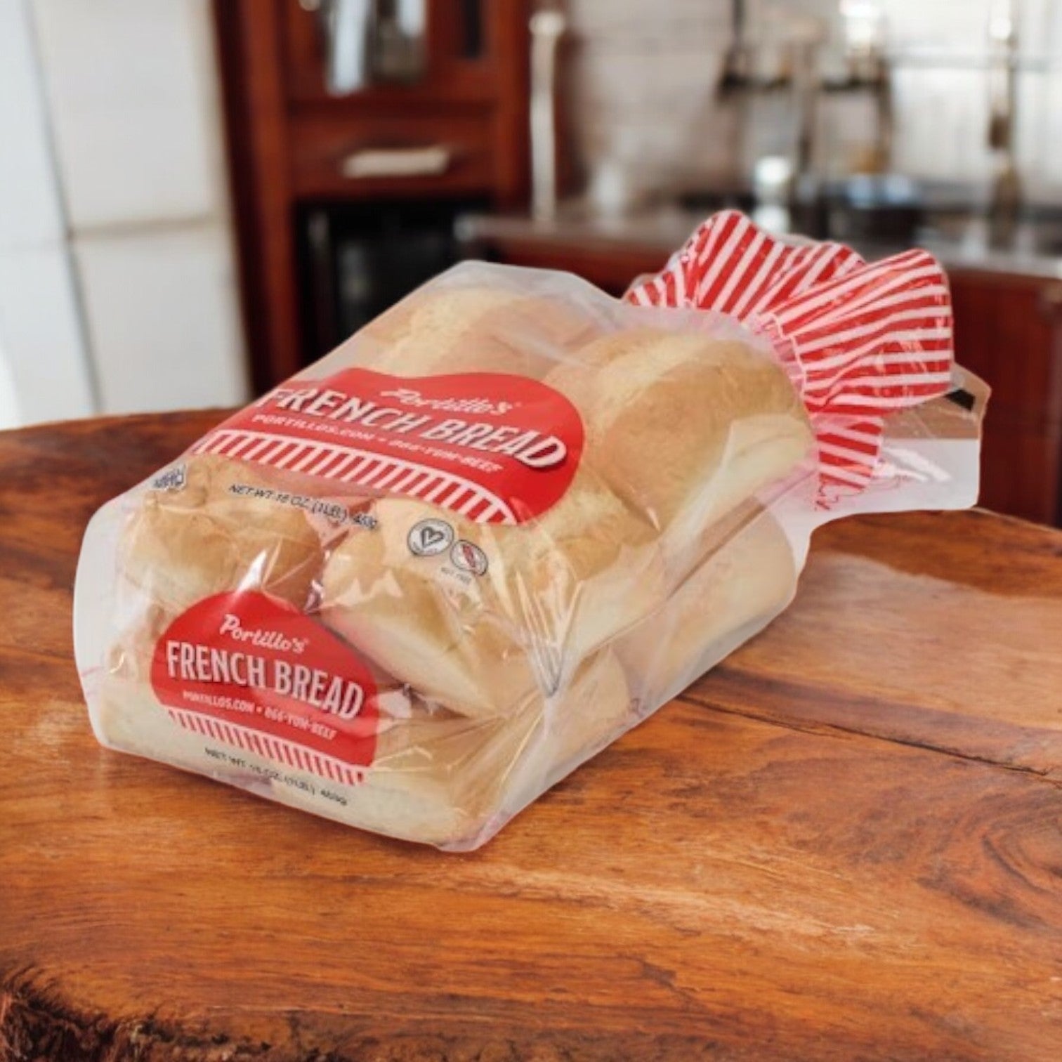 A bag of Portillo's French Bread – 16 oz, 8 Pieces 🥖 with a crispy crust sits on a wooden table in a kitchen. The bread is in clear plastic packaging featuring red and white labeling that emphasizes its freshly baked quality.