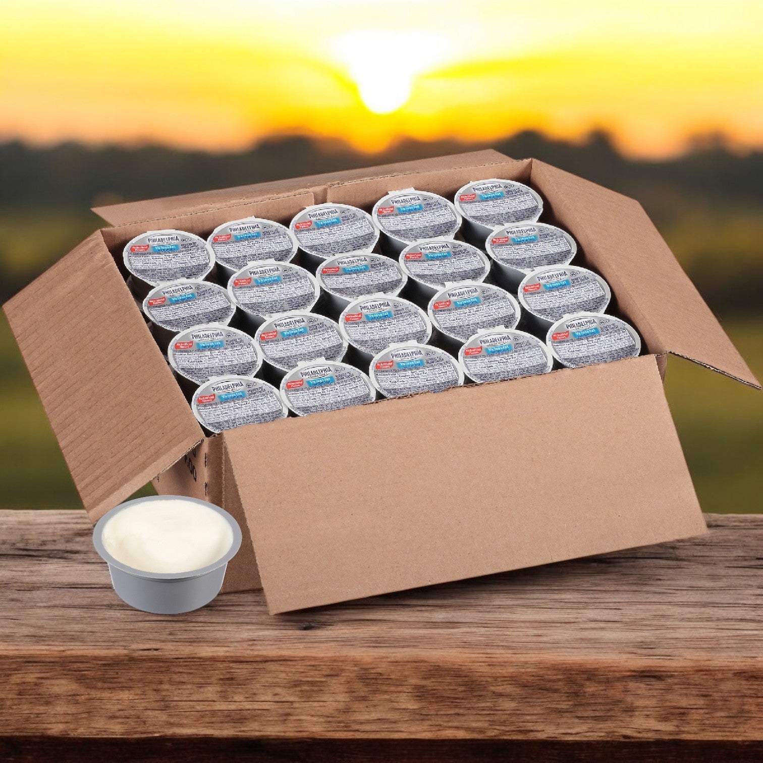 An open cardboard box filled with sealed Kraft Philadelphia Cream Cheese Light cups, each providing ideal portion control, is placed on a wooden surface against a blurred outdoor background.