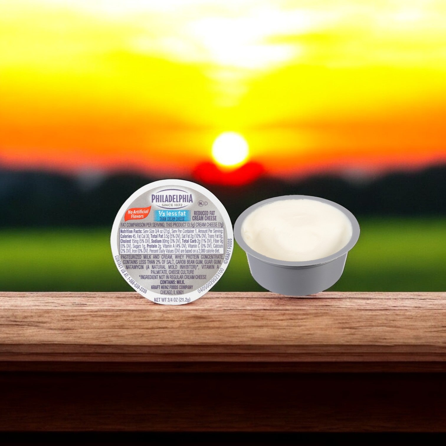 A Kraft Philadelphia Cheese Cream Light 3/4 OZ container, ideal for portion control, is beside an open cup of creamy cheese on a wooden surface, glowing warmly in the sunset.