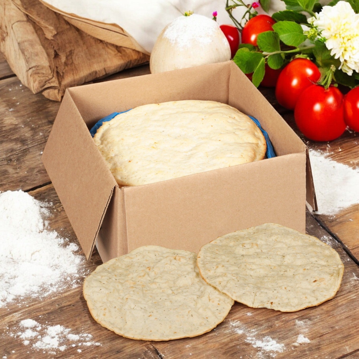 An opened box labeled "Rich’s Cauliflower Gluten-Free Pizza Crust 10", 24/5.2oz" is on a wooden table with tomatoes, flour, and a white flower scattered around. Two low-carb crusts are placed in front of the Rich's box.