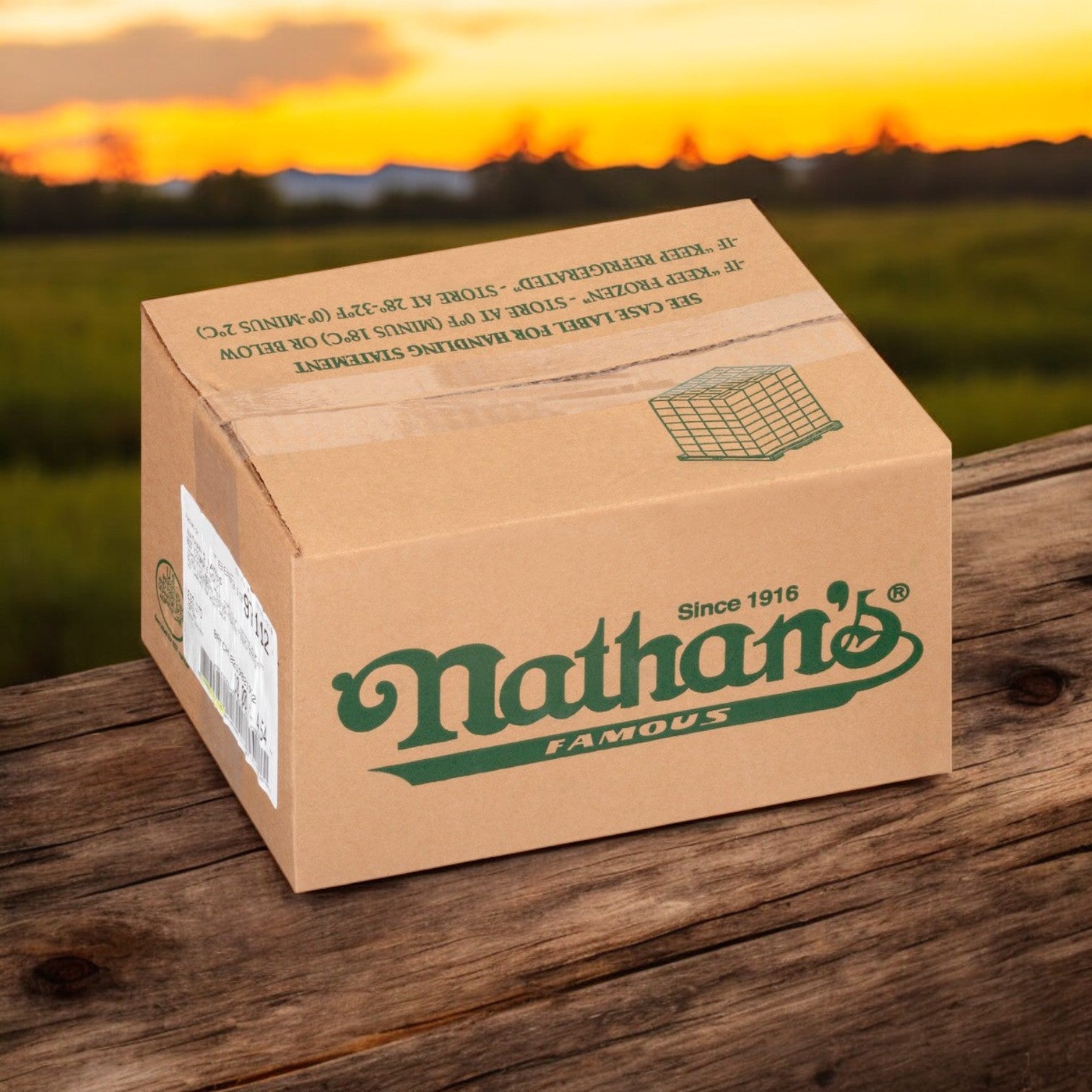 A cardboard box labeled "Nathan’s Famous Sausage Polish – 10 LB- 50 Franks In Each Case" rests on a wooden table, framed by a grassy field and brilliant sunset, evoking the rich taste of Nathan's Famous premium quality Polish sausage.