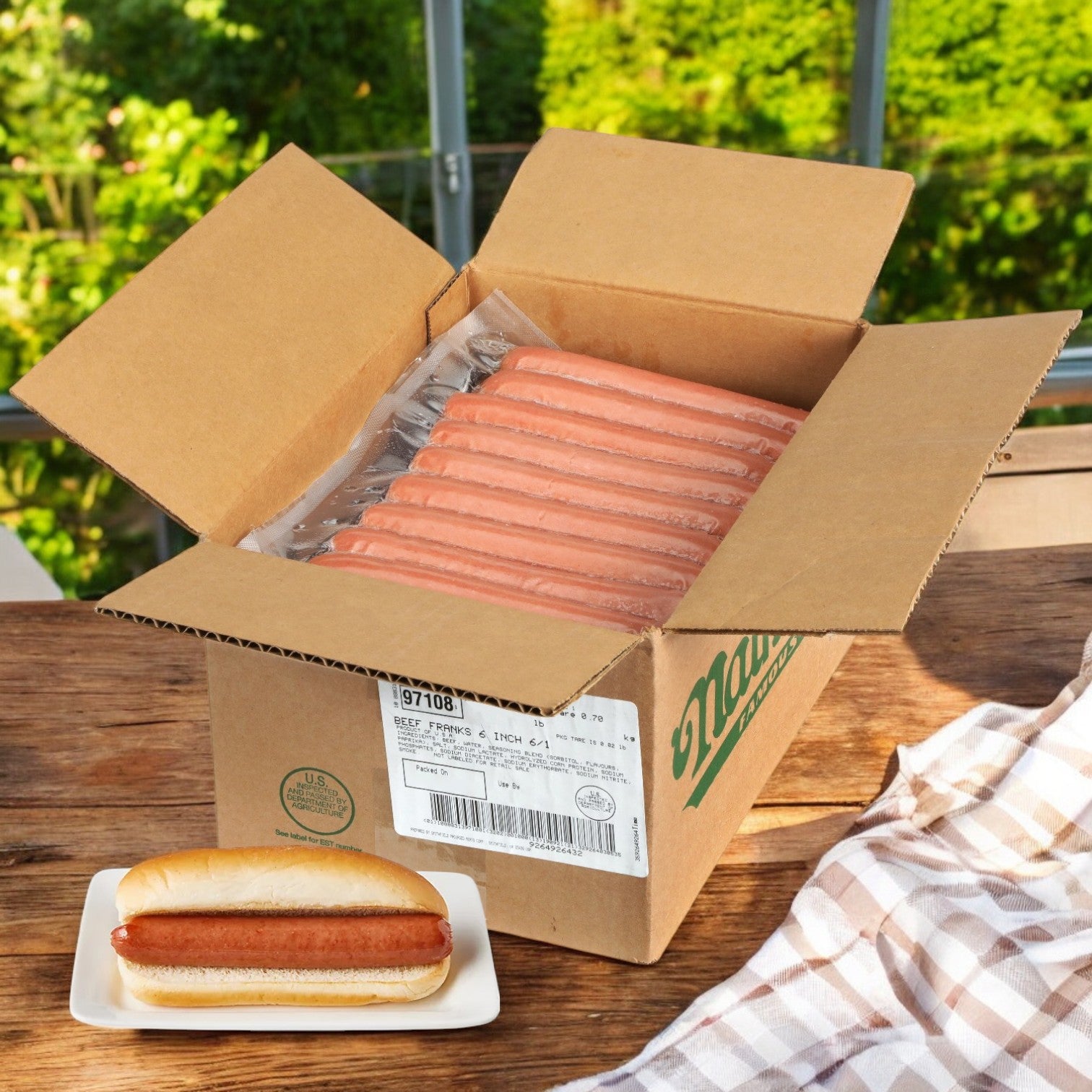 An open box of Nathan’s Famous All-Beef Frankfurters – 50 Hot Dogs sits on a wooden table beside a plate with a cooked hot dog in a bun; green foliage can be seen in the background.