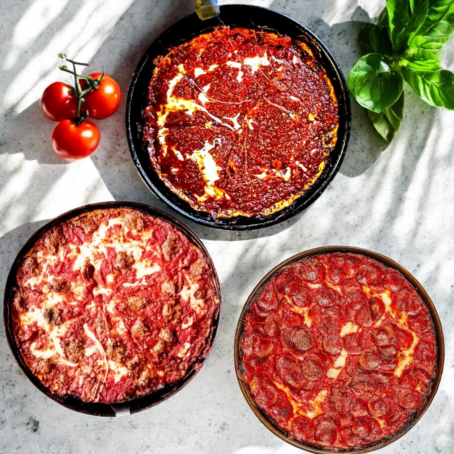 Three 10-inch deep-dish pizzas from the Pequod Pizza Deep Dish Pizza 3-Pack Combo—cheese, pepperoni, and sausage—rest on a light surface with fresh tomatoes and basil.