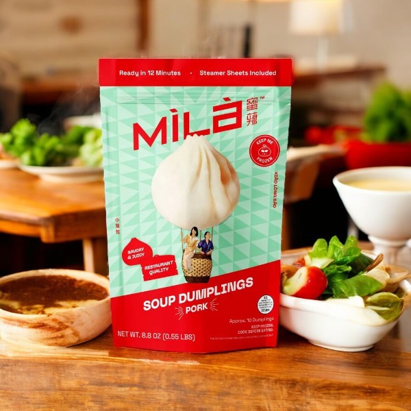 A pack of MiLa Frozen Soup Dumplings - Pork - 1 Count by Mila sits on a wooden table, next to soup, fresh greens, and vegetables, promising authentic flavor in every bite.
