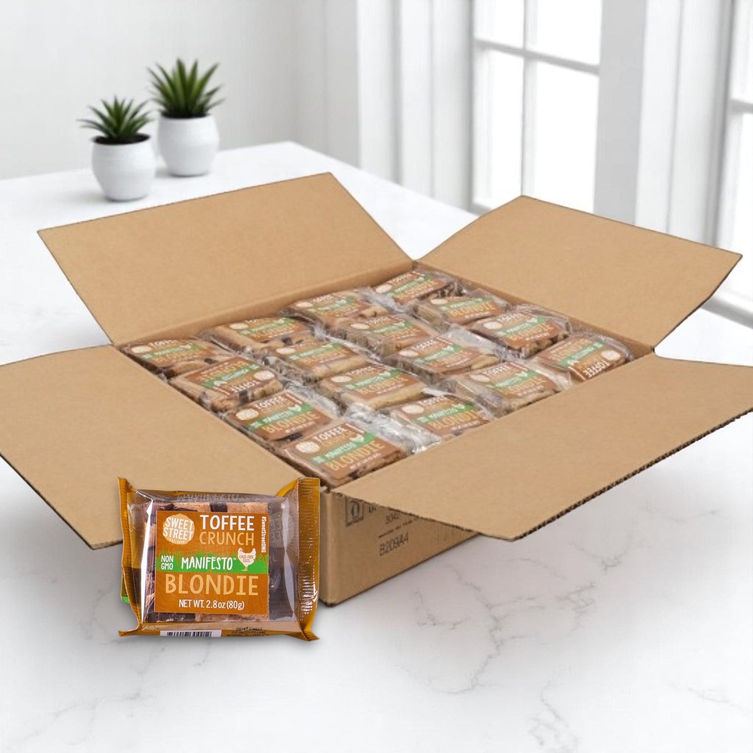 An open box of Sweet Street Blondie Toffee Brownies – 2.8 oz., 48 per case, sits on a white marble table with individually wrapped brownies, making it perfect for foodservice. Two small potted plants are in the background.