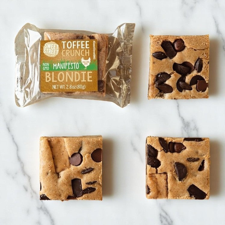 Three square Sweet Street Blondie Toffee Brownies are displayed on a marble surface beside an individually wrapped 2.8 oz. dessert labeled "Toffee Crunch Manifesto Blondie" from Sweet Street.