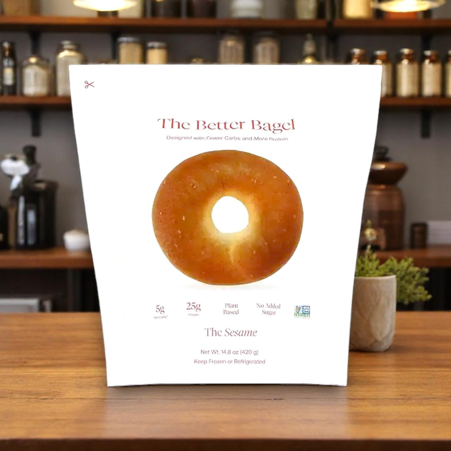 A package of BetterBrand's The Better Bagel – Sesame Style (4 bags, 16 bagels total) sits on a counter displaying its nutrition info and a photo of the high-protein bagel, with shelves holding jars and kitchen items in the background.
