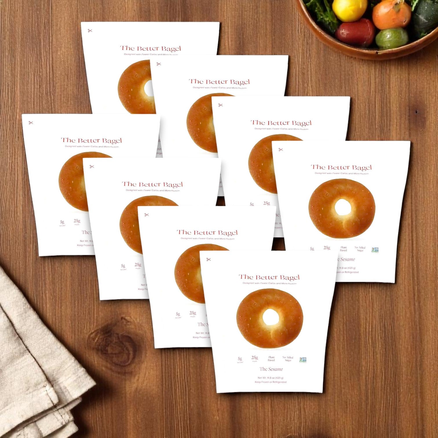 Eight bags of BetterBrand's The Better Bagel – Sesame Style (32 bagels total) are displayed on a wooden surface with a folded napkin and a bowl of cherry tomatoes. In stock and ready to ship!.
