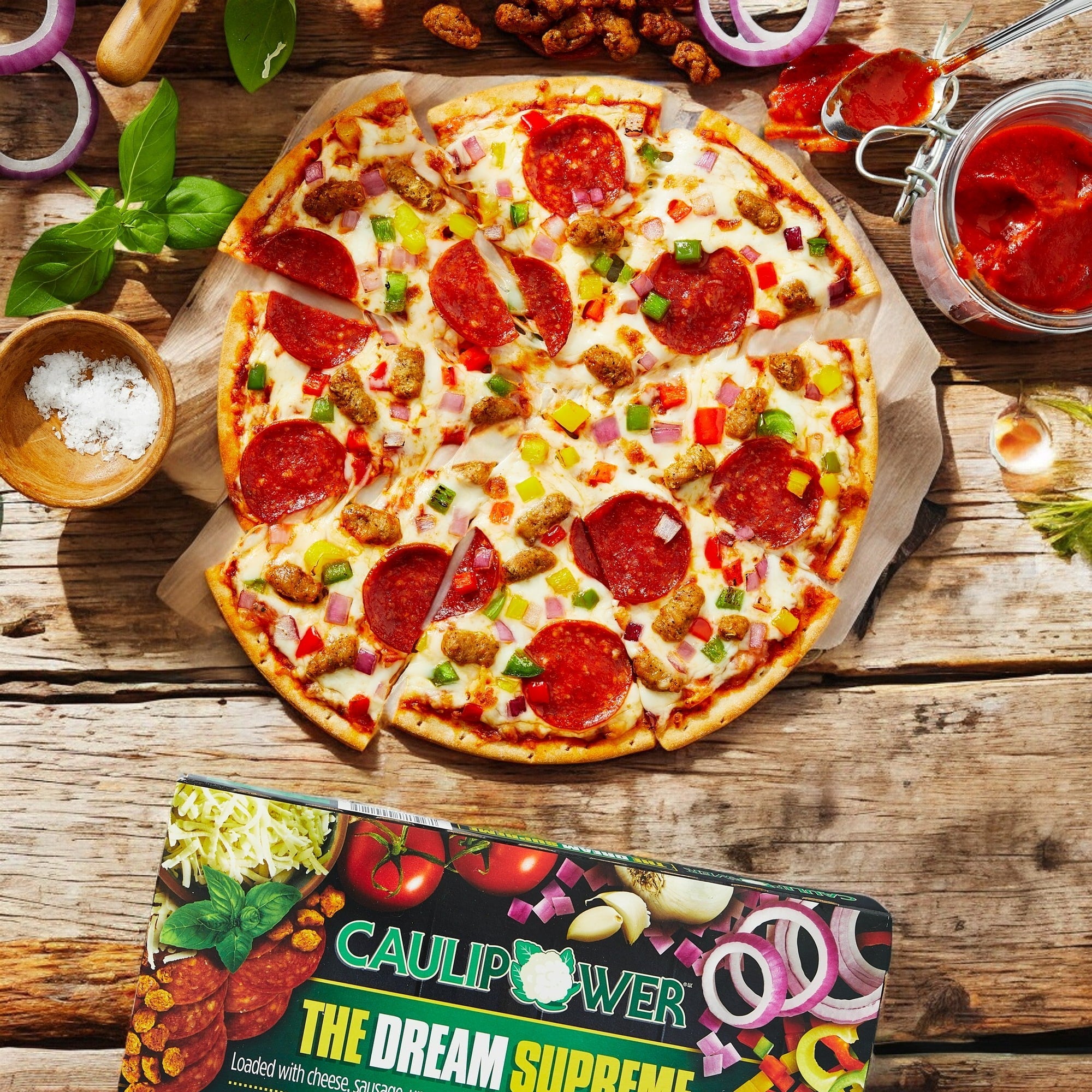 A CauliPower OTT Cauliflower Thin Crust Dream Supreme Tomato Sauce Pizza is on a wooden table, topped with pepperoni and surrounded by sliced onions, basil sprig, and tomato sauce. A partially visible box highlights the cauliflower crust option.