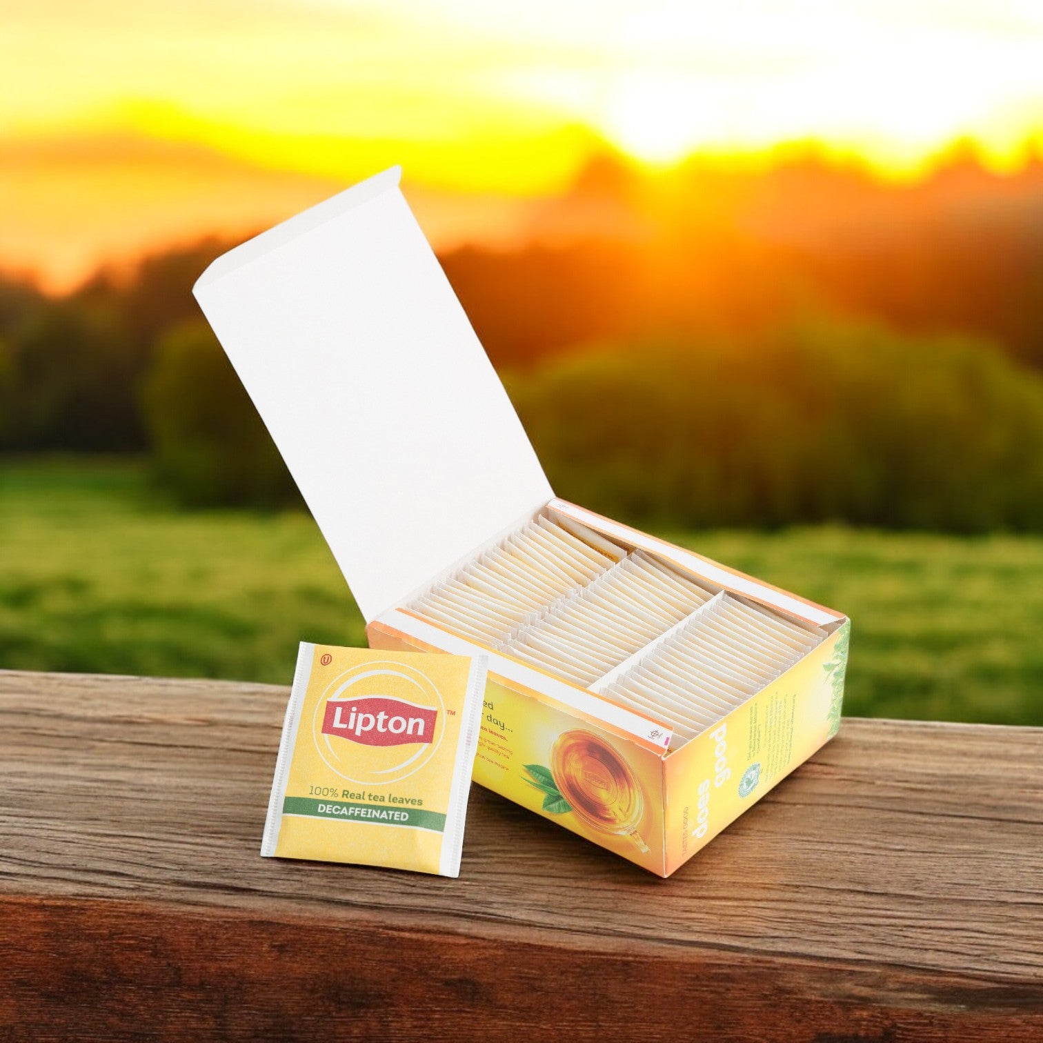 An open box of Lipton Tea Hot Bag Black Envelope Decaffeinated 6/72 CT sits on a wooden surface, its individually wrapped tea bags displayed by Lipton against a softly blurred outdoor sunset background.