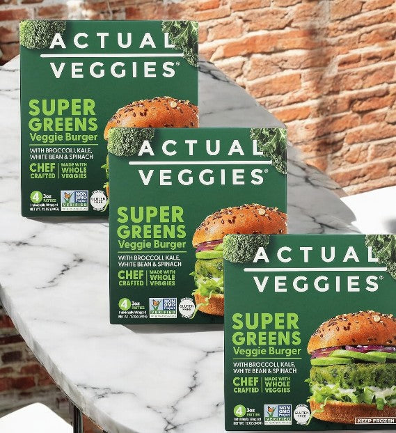 Three boxes of Actual Veggies Super Greens Veggie Burger, a plant-based patty product by Actual Veggies, are arranged on a marble surface with a brick wall background, highlighting an ideal veggie-forward meal.
