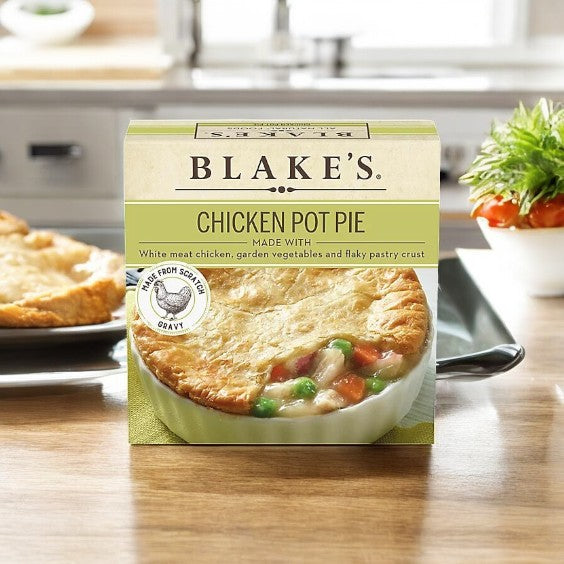 A box of Blake's All Natural Foods Chicken Pot Pie-1 Count sits on a kitchen counter, with the real pot pie and fresh vegetables in the background, showcasing this comforting frozen meal made with natural ingredients.