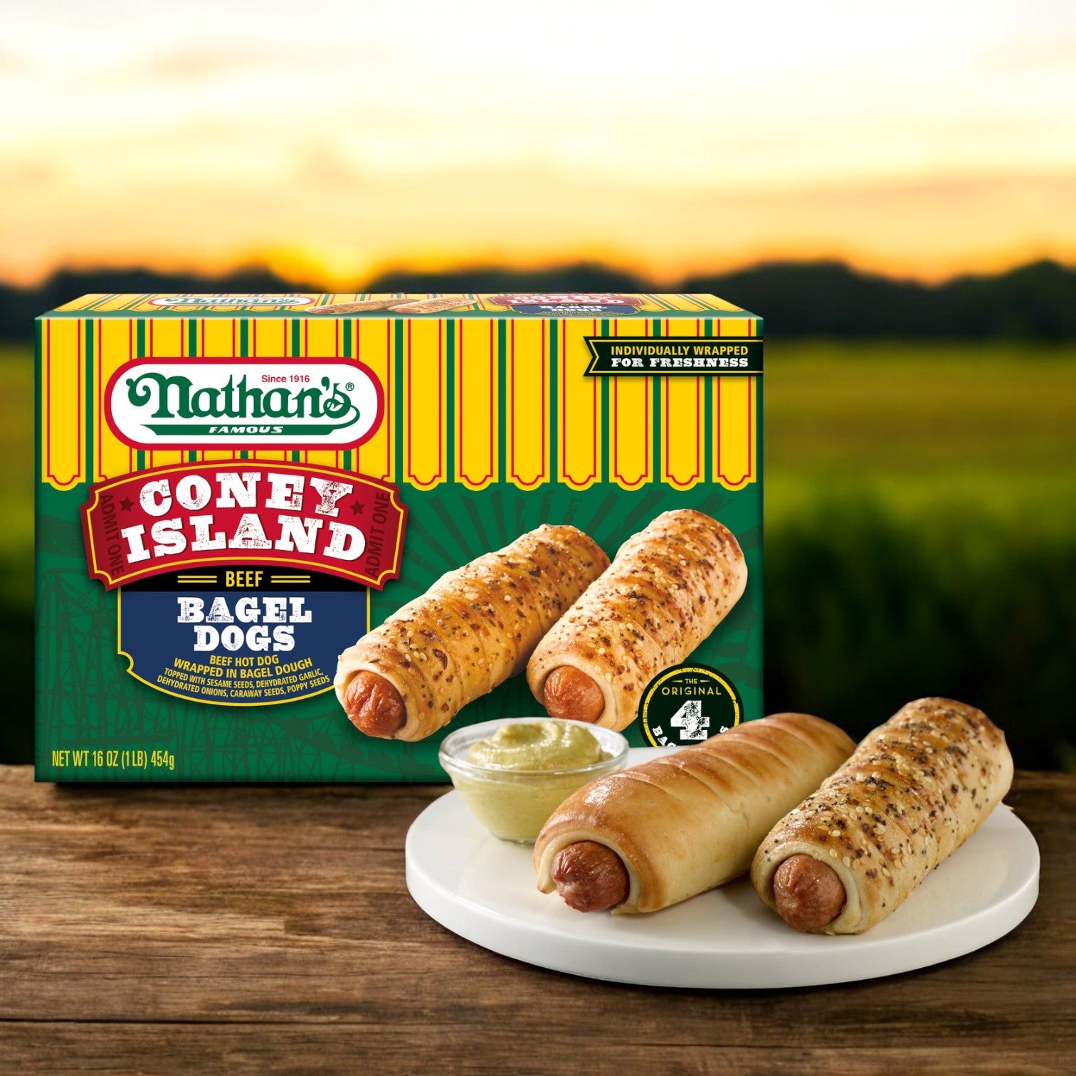 A box of Nathan's Famous Coney Island Bagel Dogs – 20 oz. (4 Dogs per Pack) sits behind a plate with two premium beef bagel dogs and a small dish of mustard, all set on an outdoor table.