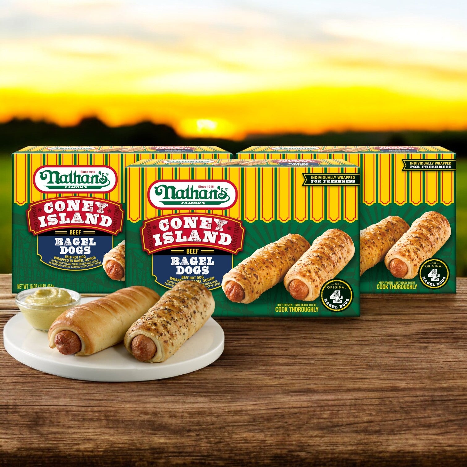 Three boxes of Nathan’s Famous Coney Island Bagel Dogs – 3-Pack (4 Dogs per 20 oz. Pack) by Nathan’s Famous are shown behind a plate with two bagel-wrapped premium beef hot dogs and mustard, on a wooden table at sunset.