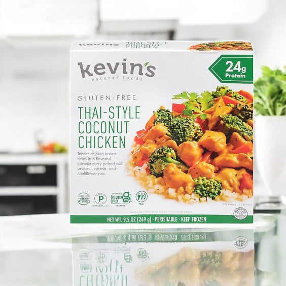 A box of Kevin's Thai-Style Coconut Chicken - 1 Count, labeled gluten-free, paleo-friendly, and 24g protein, sits on a kitchen counter with the tasty meal shown on the front.